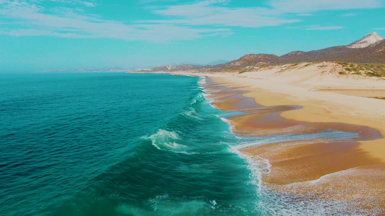 Aerial view over Punta Buzzards in Baja California Sur with turquoise ocean at left, a single breaking wave near shore, broad sand, dunes, distant hills, and no visible people.