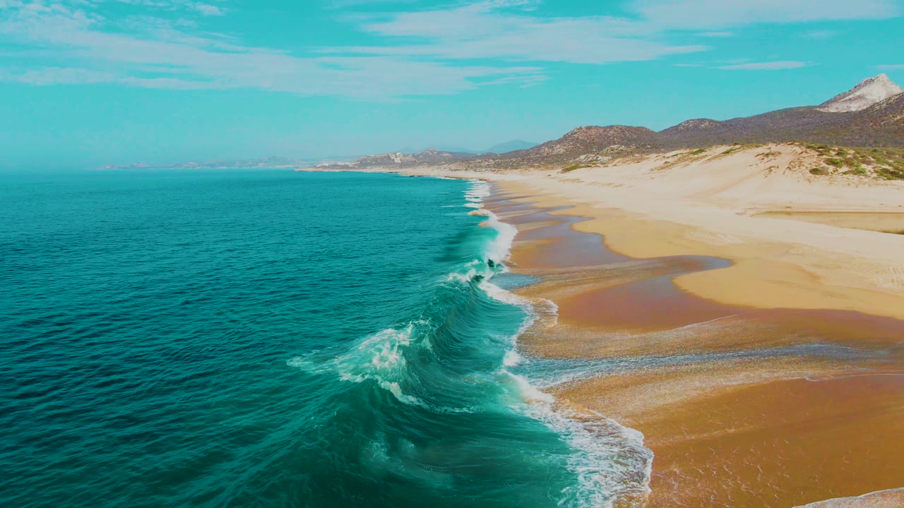 Aerial frame from Punta Buzzards showing a curling turquoise wave breaking beside wet sand pools, with pale dunes and low hills in the background and no visible people.
