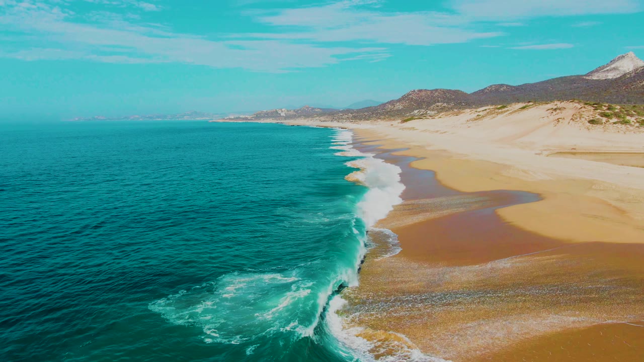 Punta Buzzards aerial view with white foam spreading from a breaking wave across the nearshore water, tan beach to the right, coastal hills beyond, and no visible people.