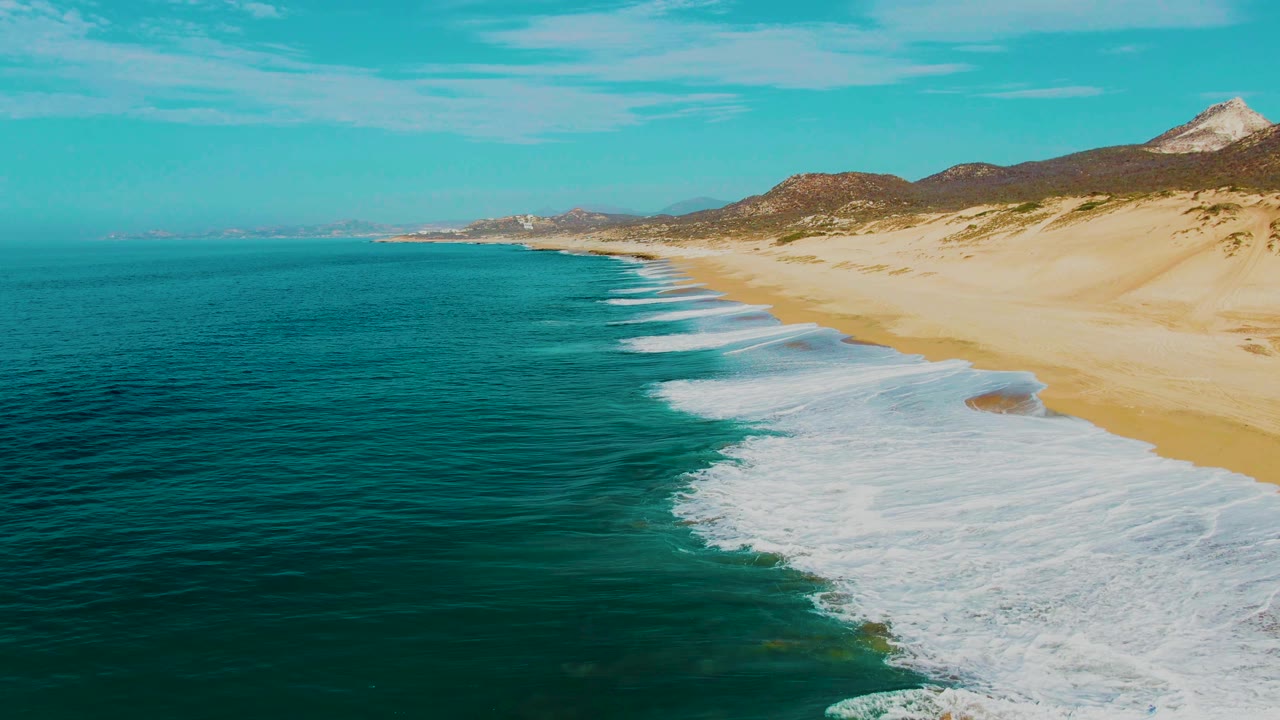 Aerial coastline with turquoise ocean, white surf, sandy beach, and distant hills.