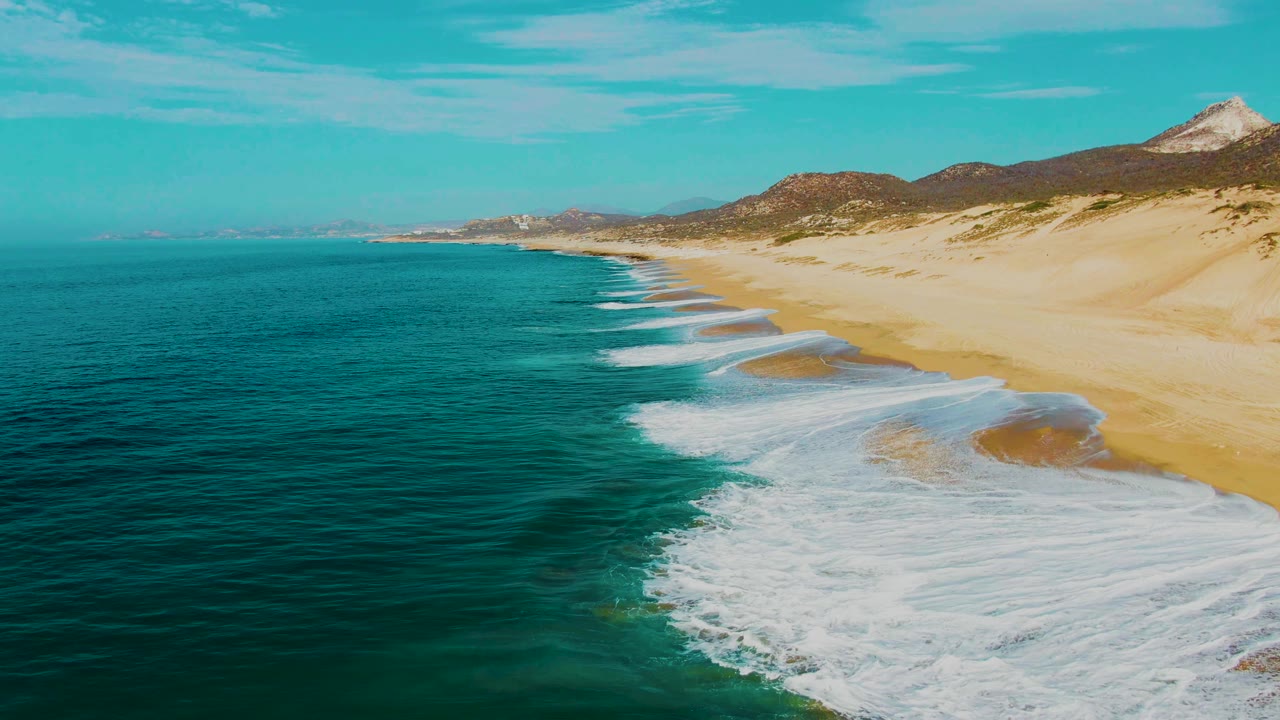 Aerial frame at Punta Buzzards showing more foam bands along the shoreline, deep turquoise water to the left, broad sand to the right, coastal hills in the background, and no visible people.