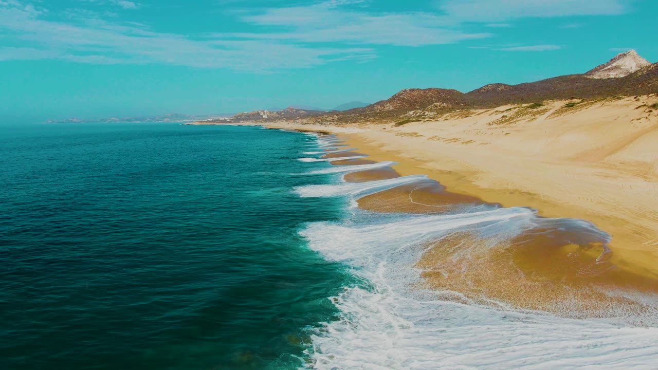 Aerial view over Buzzards Beach with a larger white foam wash crossing wet sand, turquoise ocean on the left, pale dunes and hills beyond the beach, and no visible people.