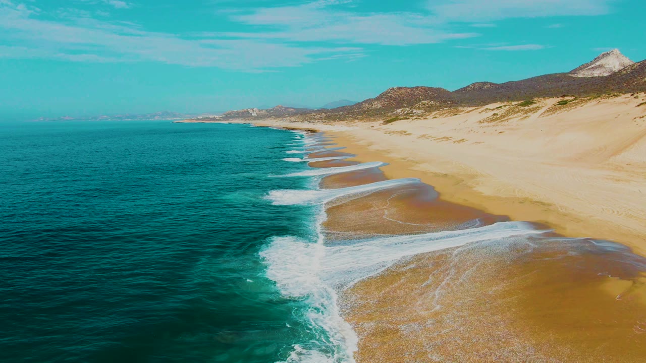Aerial frame of the East Cape shoreline with a long diagonal beach, surf curling in from the lower left, turquoise water offshore, hills along the horizon, and no visible people.