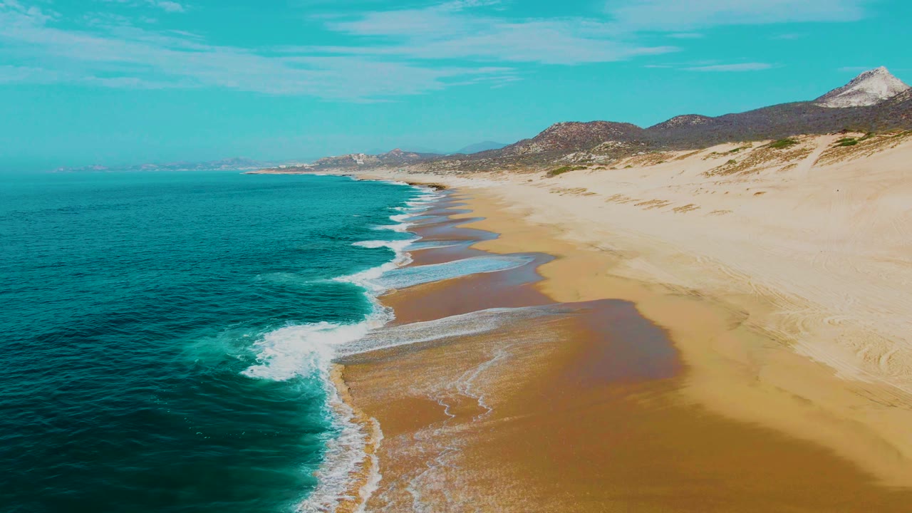 Aerial view of turquoise ocean meeting a sandy beach with distant hills.