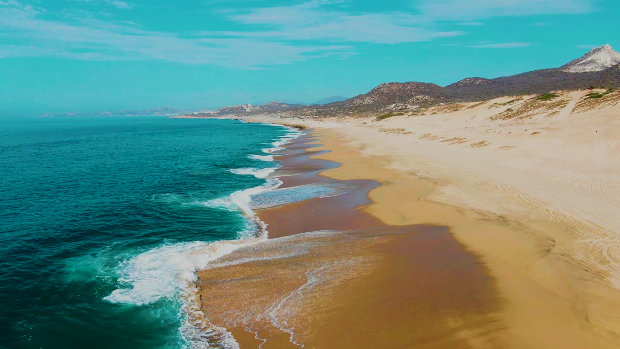 Aerial frame from Punta Buzzards, Baja California Sur with a thicker band of white foam reaching the beach, turquoise ocean to the left, sand flats to the right, and no visible people.