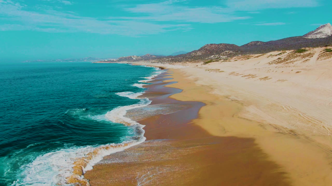 Aerial frame from Punta Buzzards, Baja California Sur where the foamy surf curves diagonally across wet sand, with open ocean at left and no visible people.