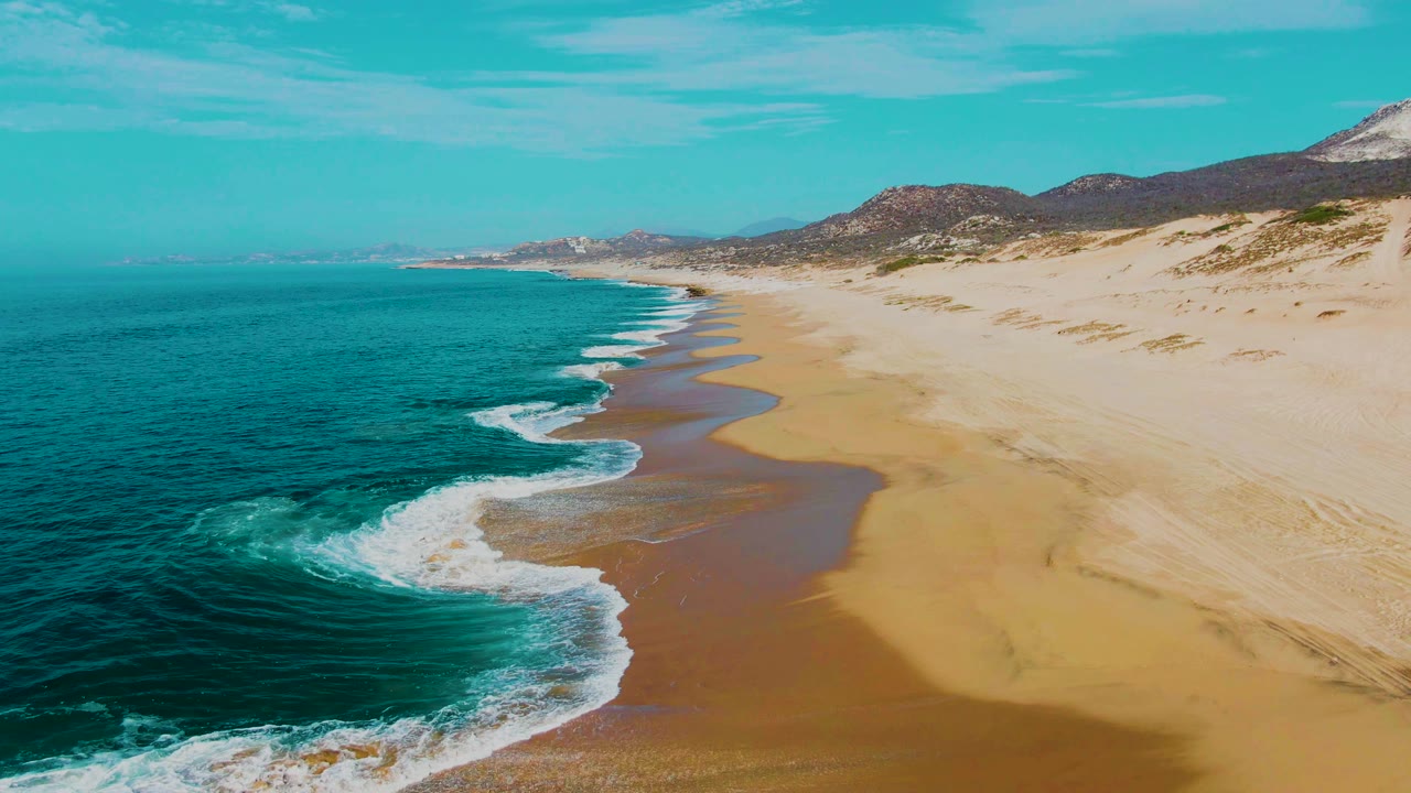Aerial frame from Punta Buzzards, Baja California Sur showing an empty sandy shoreline, a scalloped edge of white surf, turquoise water, low dunes, distant hills, and no visible people.