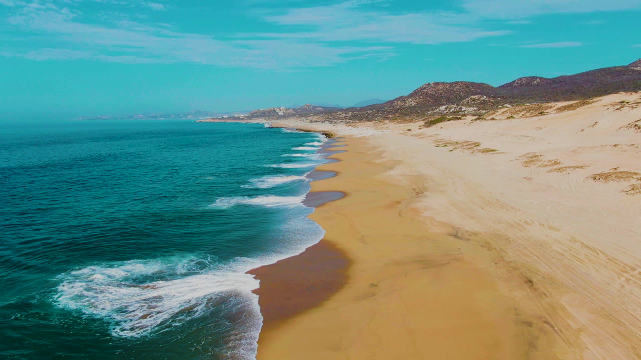 Aerial frame at Punta Buzzards, Baja California Sur, showing turquoise ocean on the left, white surf touching golden sand, low hills beyond the beach, and no visible people.