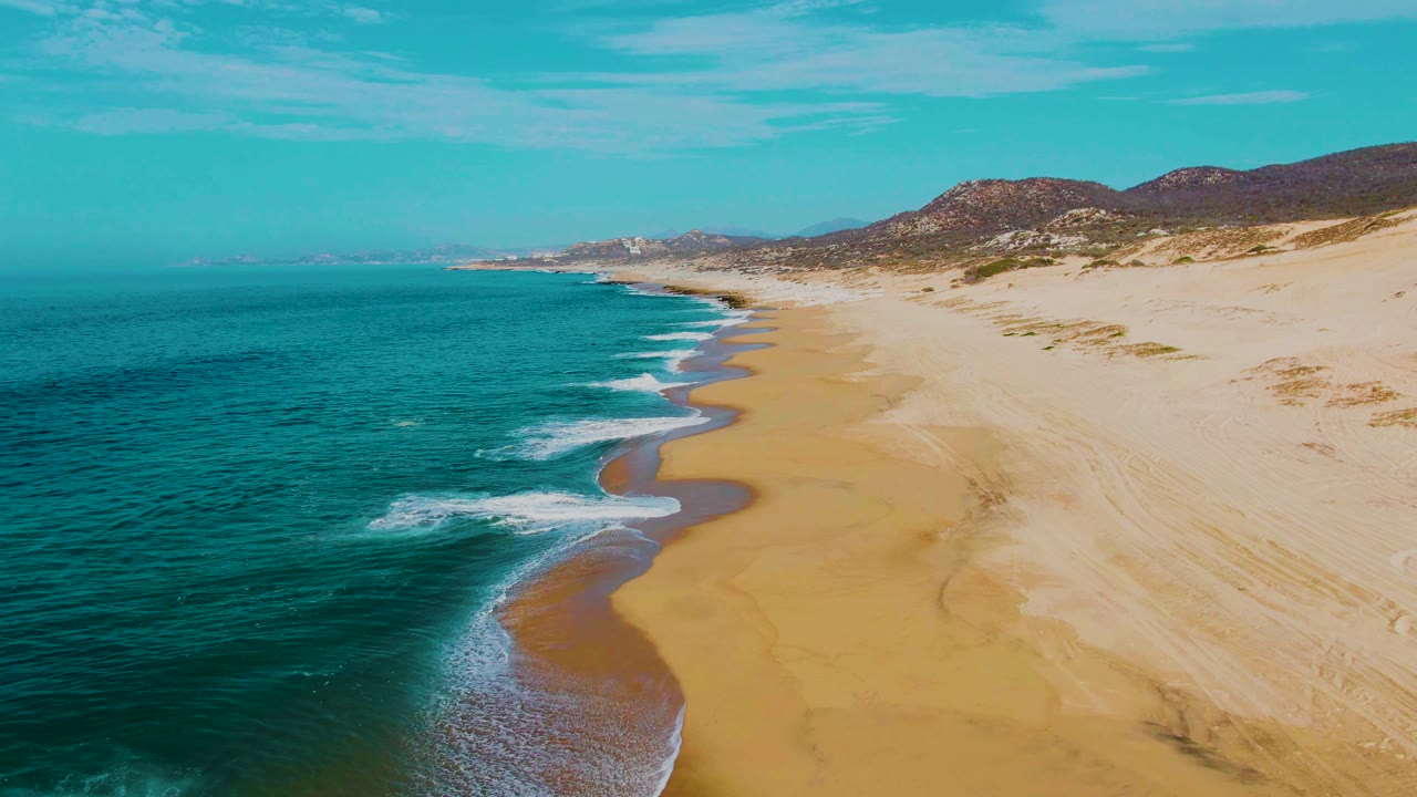 Aerial frame at Punta Buzzards, Baja California Sur, where a larger foamy wave breaks near the lower shoreline while the beach and dune-backed hills recede into the distance, with no visible people.