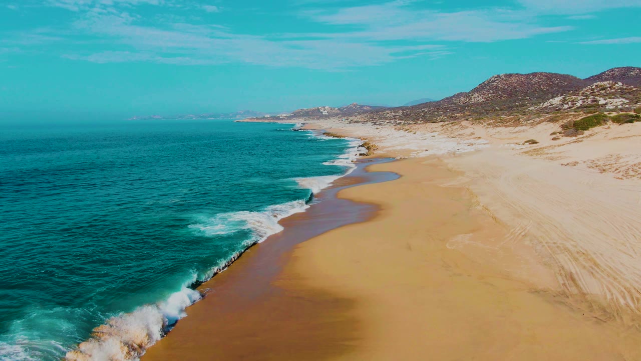 Aerial view over Punta Buzzards in Baja California Sur, with turquoise water on the left, white foam at the shoreline, wide sand on the right, distant hills, and no visible people.