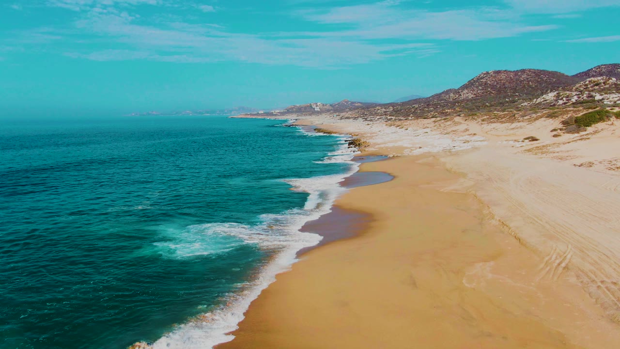 Turquoise surf washes farther along a wide sandy beach under a clear sky.