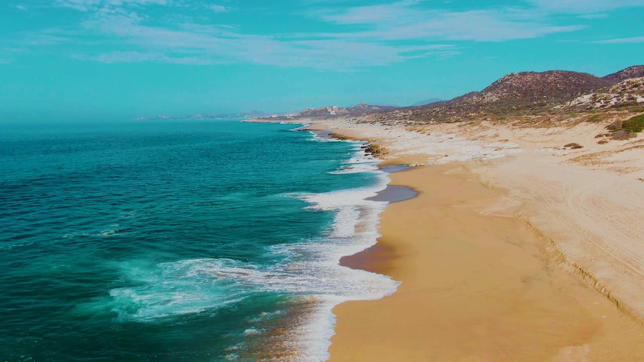 Aerial view at Punta Buzzards with white breakers spreading across the shoreline, deeper turquoise water offshore, pale sand to the right, distant hills, and no visible people.