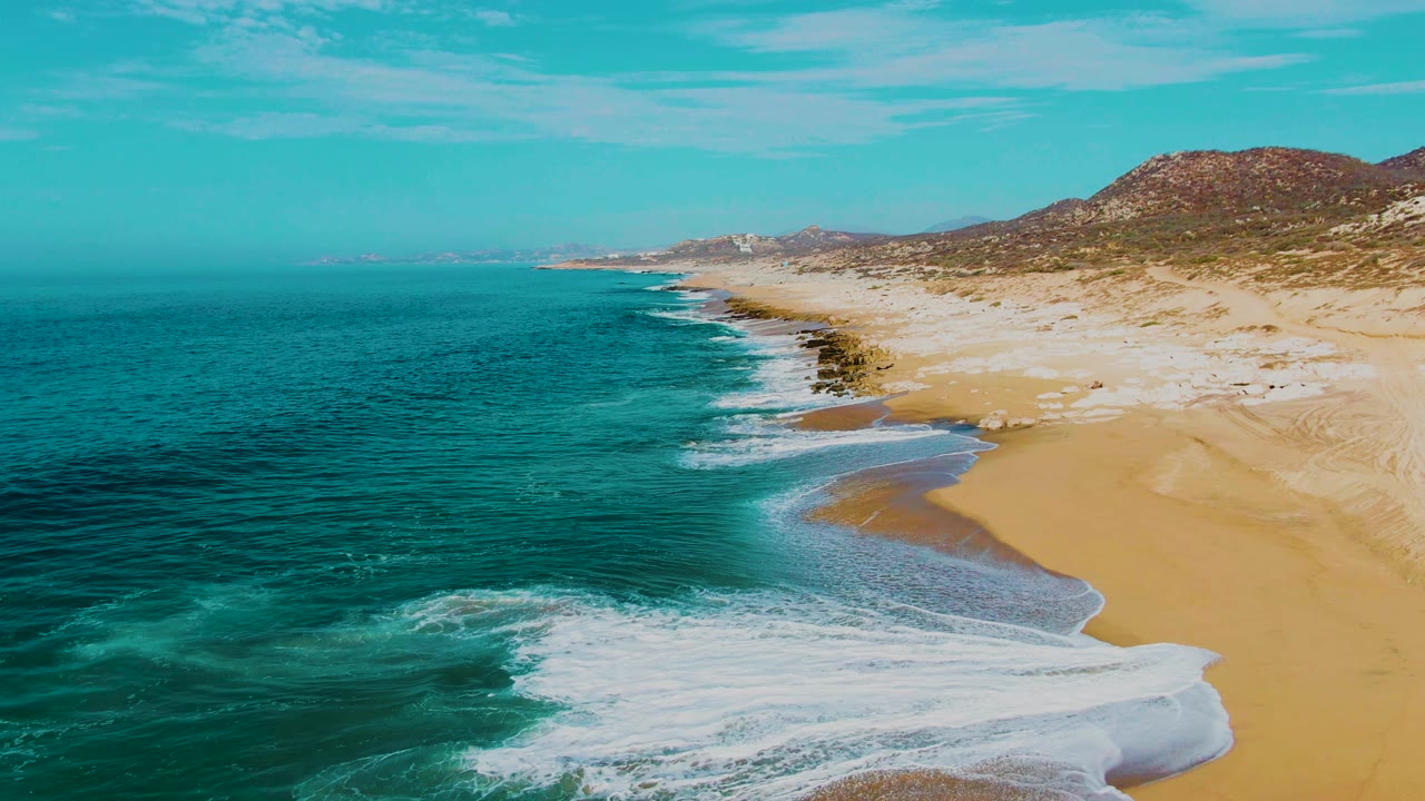 Aerial view of turquoise ocean waves reaching a sandy coastline with low hills inland.