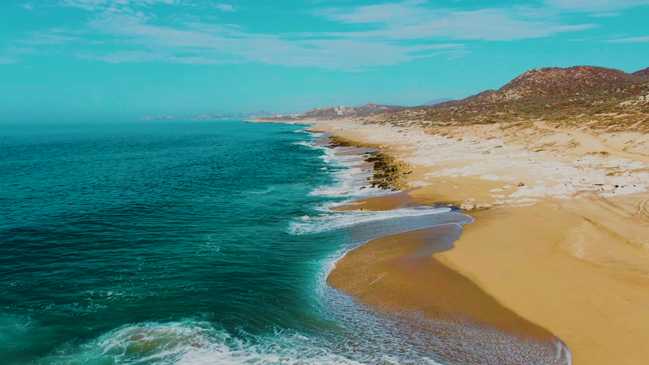 Aerial frame from Punta Buzzards showing a curved wash of foam crossing wet sand, dark shoreline rocks, turquoise water, low hills, and no visible people.