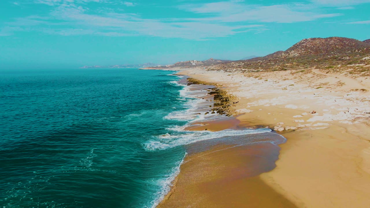 Drone-style aerial view at Punta Buzzards with more beach visible on the right, surf breaking along dark rocks, turquoise water to the left, and no visible people.