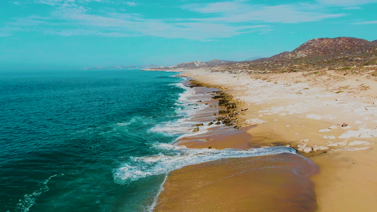 Mid-clip aerial frame over Buzzards Beach, Baja California Sur, where white water wraps around shoreline rocks and shallow sand patches with no visible people.