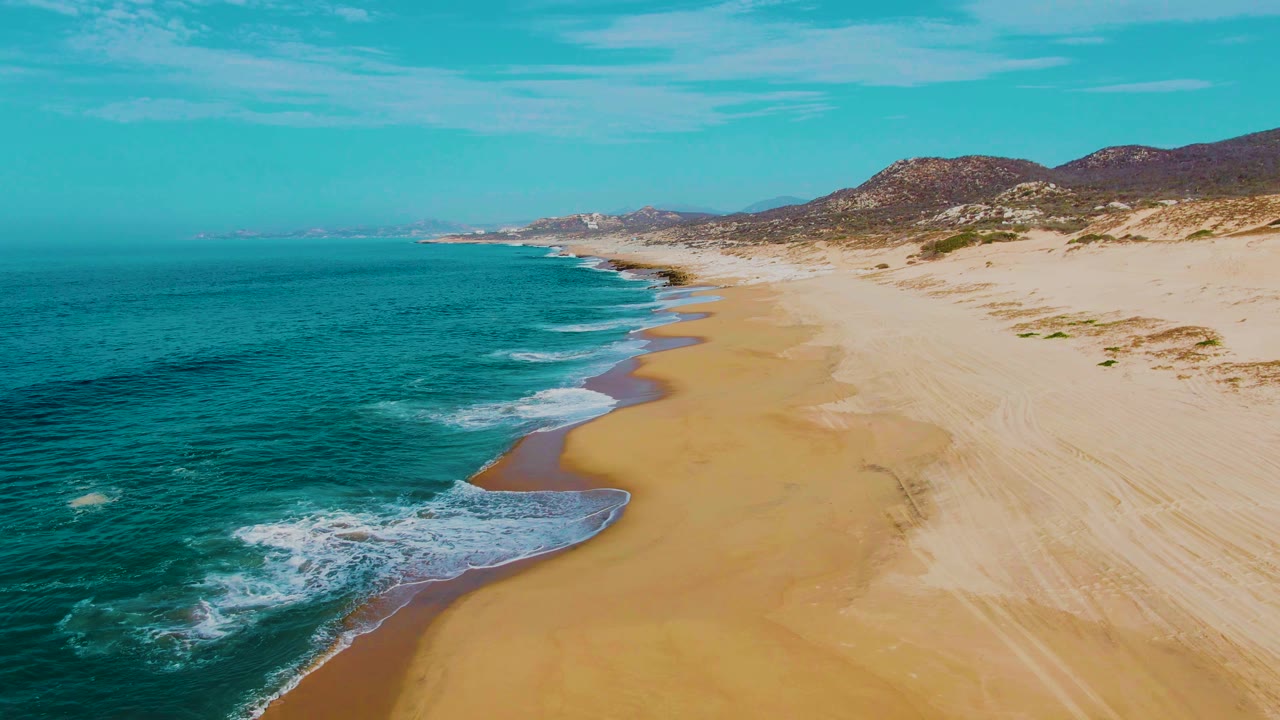 Aerial view of turquoise ocean beside a sandy beach and dunes.