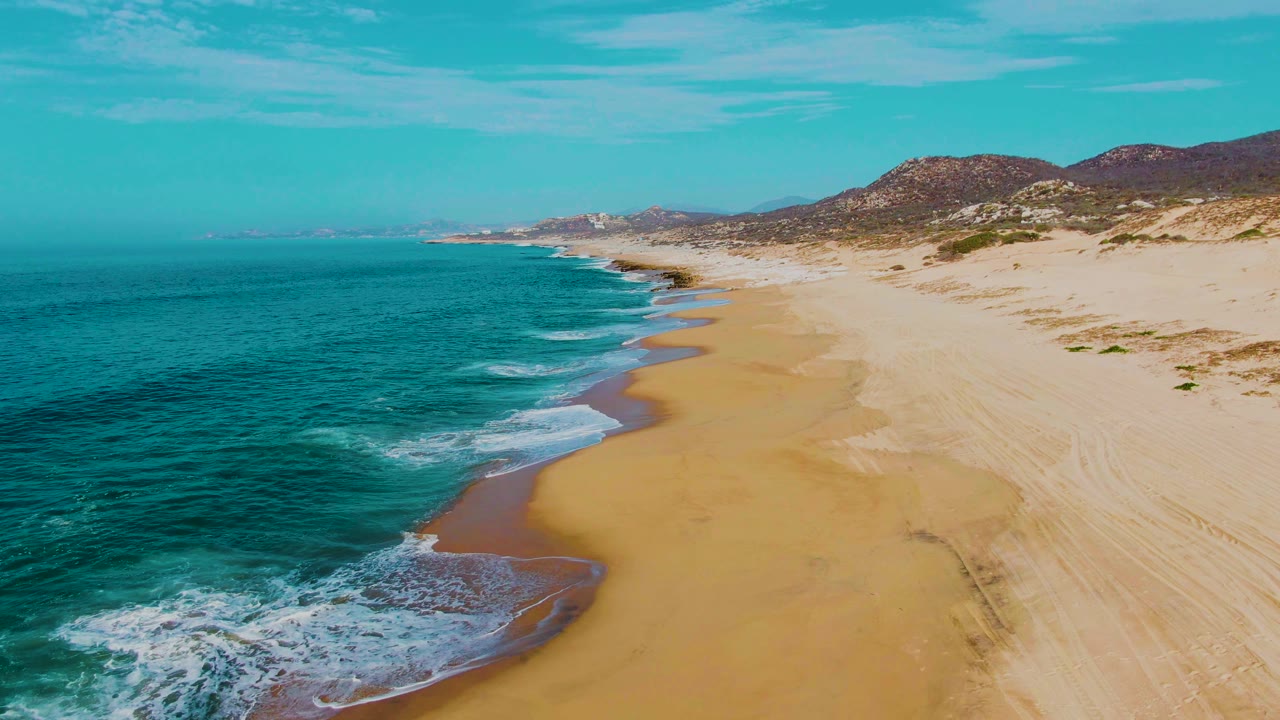 Aerial frame over Punta Buzzards with white foam bands moving toward wet sand, dry dune tracks on the right, low hills in the distance, and no visible people.