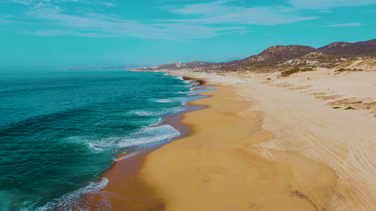 Punta Buzzards beach seen from above with a darker turquoise sea at left, a smooth tan shoreline curving through center, dune ridges at right, and no visible people.