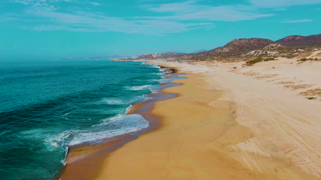 High coastal view at Punta Buzzards where a white surf patch reaches the lower left shoreline, open sand stretches across the middle, dunes sit to the right, and no visible people appear.