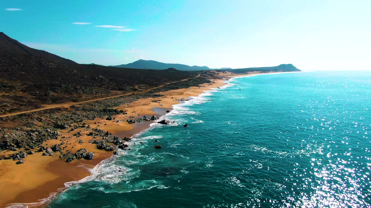 Aerial view over Punta Buzzards with turquoise ocean on the right, waves breaking onto a rocky sandy beach, dark hills inland, a coastal road, and no visible people.