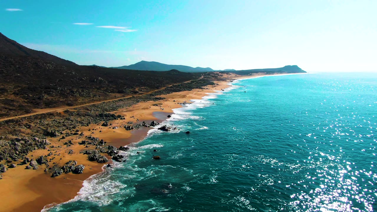 Wide aerial frame at Punta Buzzards showing bright sun glints on turquoise water, foamy waves meeting scattered rocks and sand, inland hills, and no visible people.