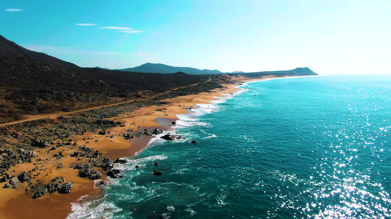 Aerial view of the Punta Buzzards shoreline with a cluster of rocks near the lower beach, white surf washing around them, open turquoise sea, distant hills, and no visible people.