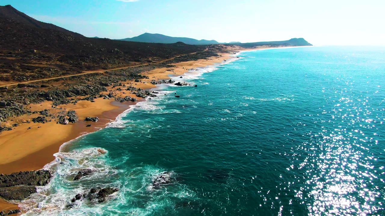 Aerial view along Punta Buzzards beach with bright turquoise water to the right, white foam at the shoreline, rocky sand in the foreground, distant mountains, and no visible people.