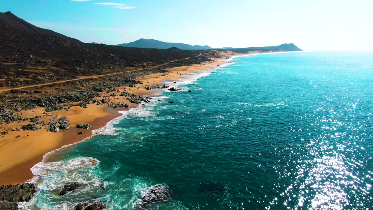 Aerial frame of Punta Buzzards with sun glitter across the ocean, a rocky sandy beach curving through the left side, low mountains inland, and no visible people.