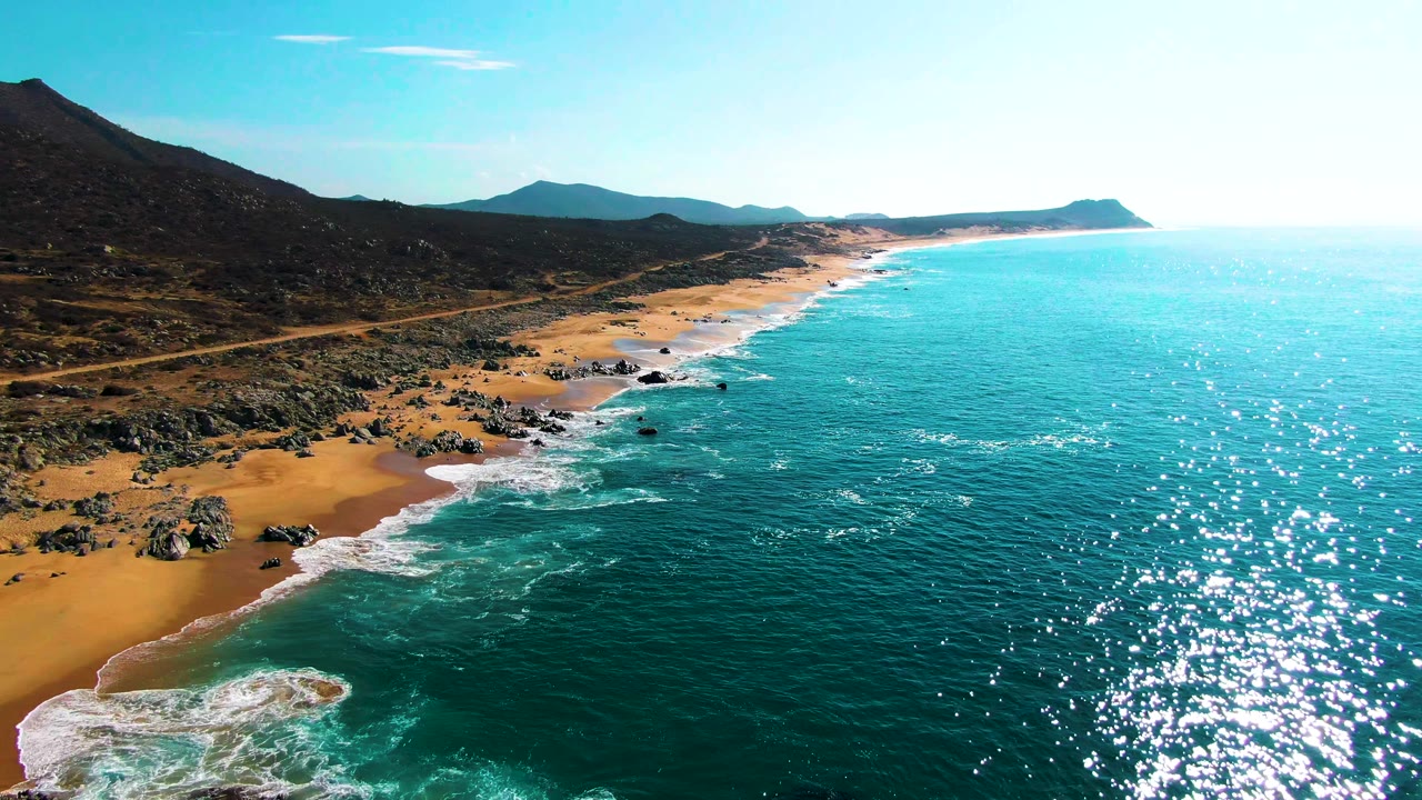 Wide aerial frame over the Punta Buzzards coast where white surf breaks beside clustered rocks and golden sand, with a road and mountain slopes inland and no visible people.