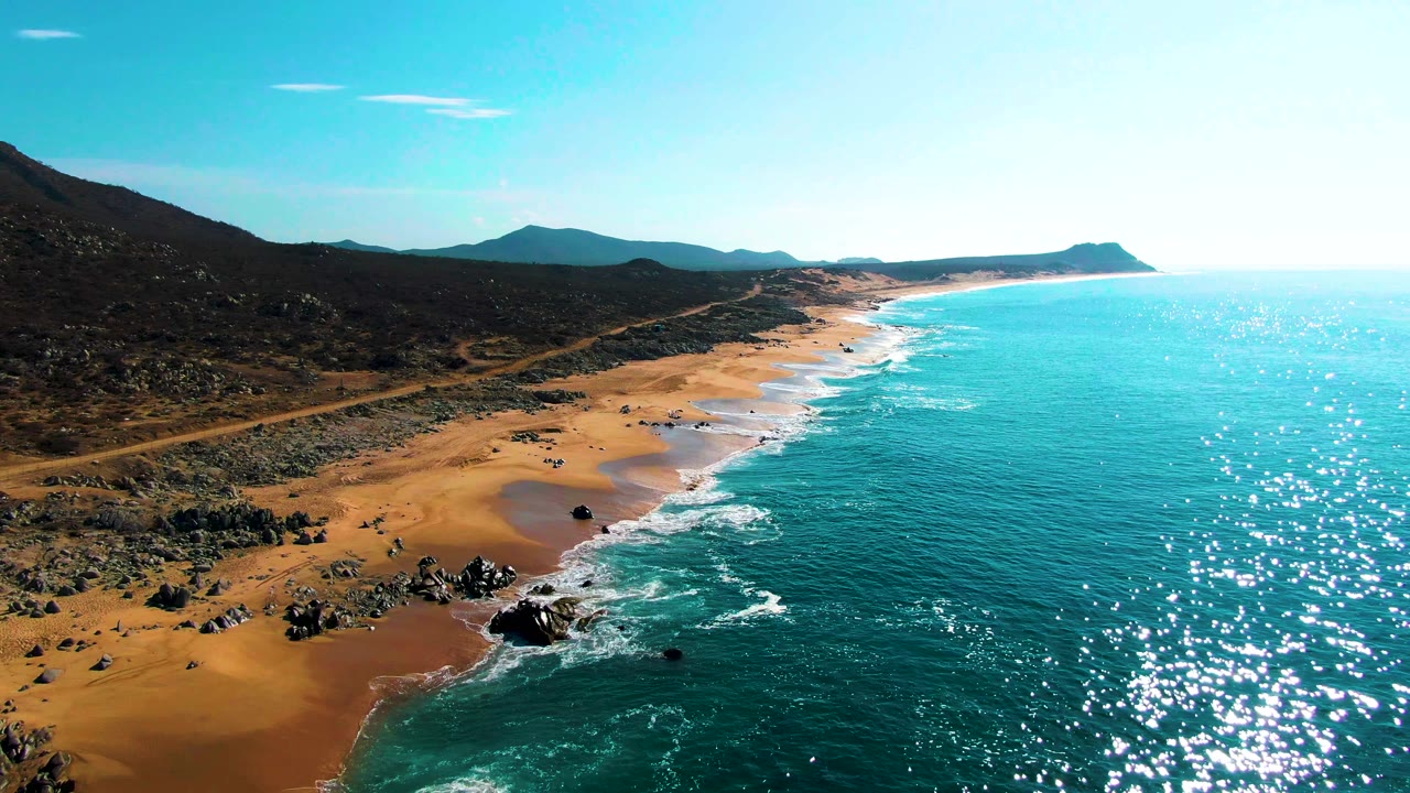 Drone-height view along Buzzards Beach with foamy waves tracing the sand, dark rocky terrain inland, a long shoreline curve, distant mountains, and no visible people.