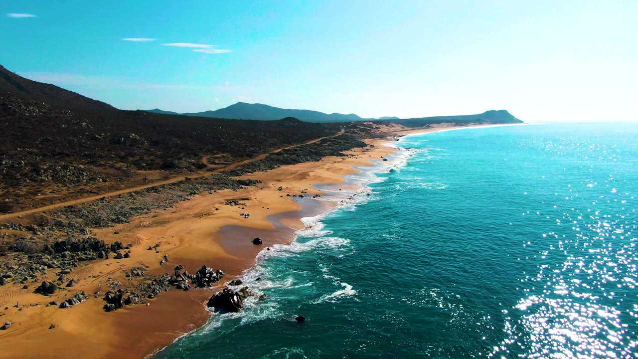 Aerial frame from Punta Buzzards showing sun sparkle across turquoise water, a rocky section of sandy beach in the lower left, a curving coast ahead, mountains inland, and no visible people.