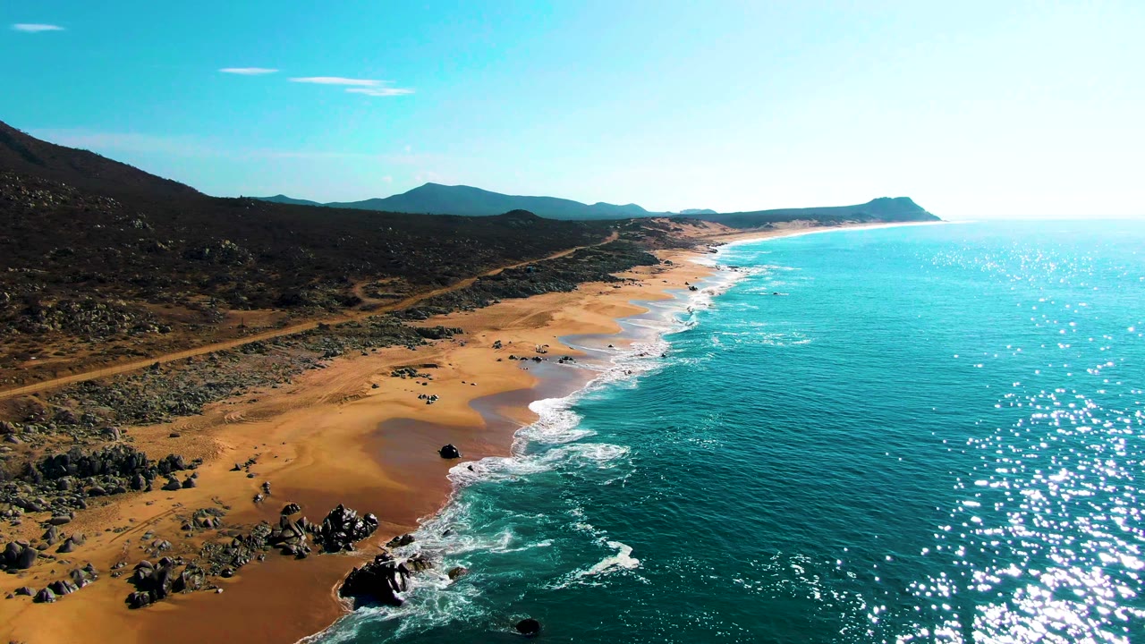 High aerial view of Buzzards Beach with rocky outcrops in the lower left foreground, turquoise surf rolling onto sand, dark inland hills, a distant headland, and no visible people.