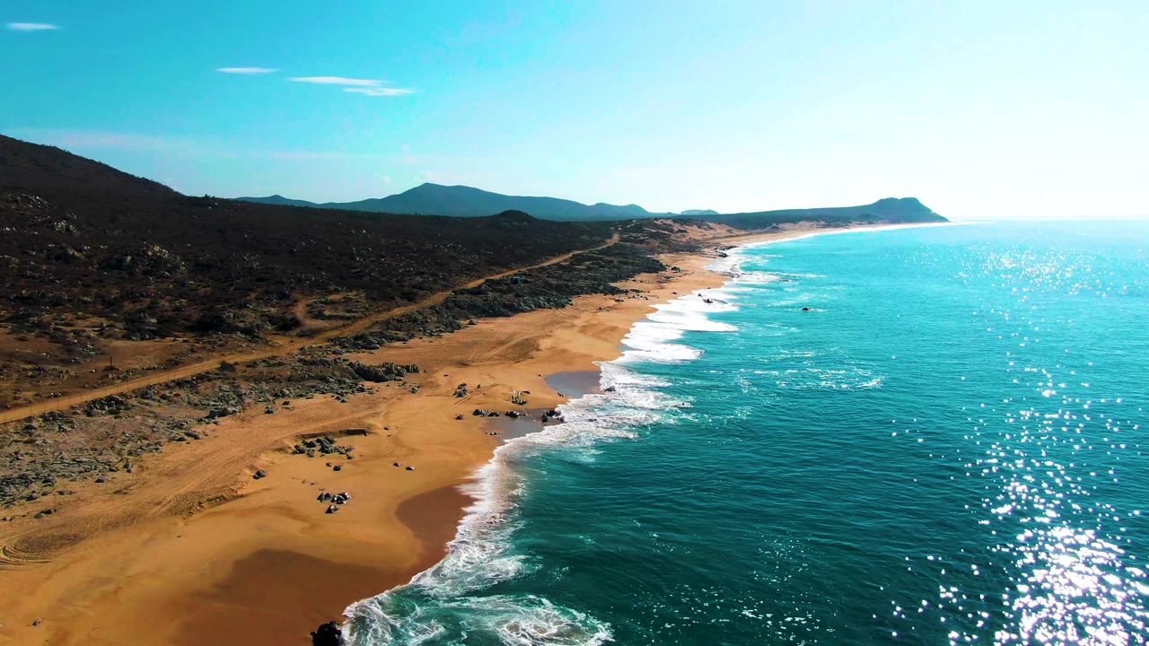 Aerial view over Buzzards Beach in Baja California Sur, with turquoise water on the right, surf reaching tan sand, rocky terrain inland, distant mountains, and no visible people.