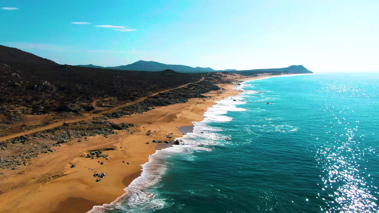 At 0:01, the Buzzards Beach shoreline extends farther into the distance, with bright sun glitter on the turquoise sea, white wave bands on the sand, inland rocks, and no visible people.