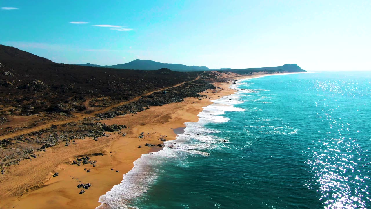 At 0:02, the aerial angle frames Buzzards Beach with a wide strip of tan sand, repeated white surf lines, turquoise water, rocky desert terrain, distant mountains, and no visible people.