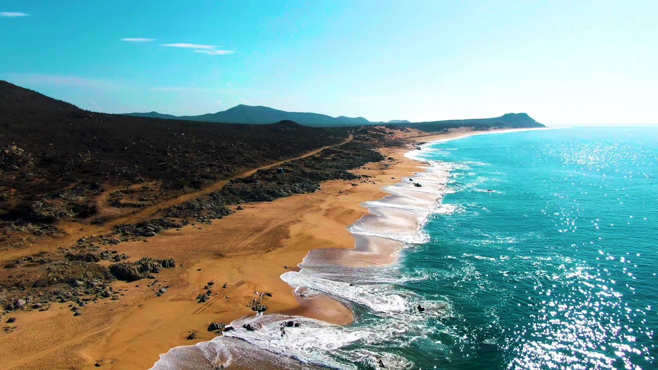 Aerial view over Buzzards Beach in Baja California Sur, with turquoise water glittering beside a sandy shoreline, dark rocky scrubland inland, distant hills, and no visible people.