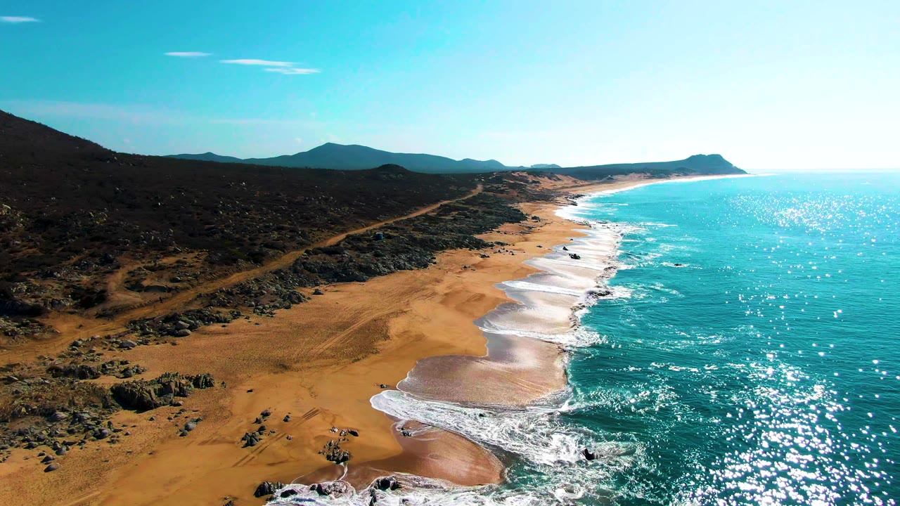 Drone perspective along Buzzards Beach, Baja California Sur, showing white wave lines curling onto wet sand, a dirt track through rocky coastal scrub, distant mountains, and no visible people.