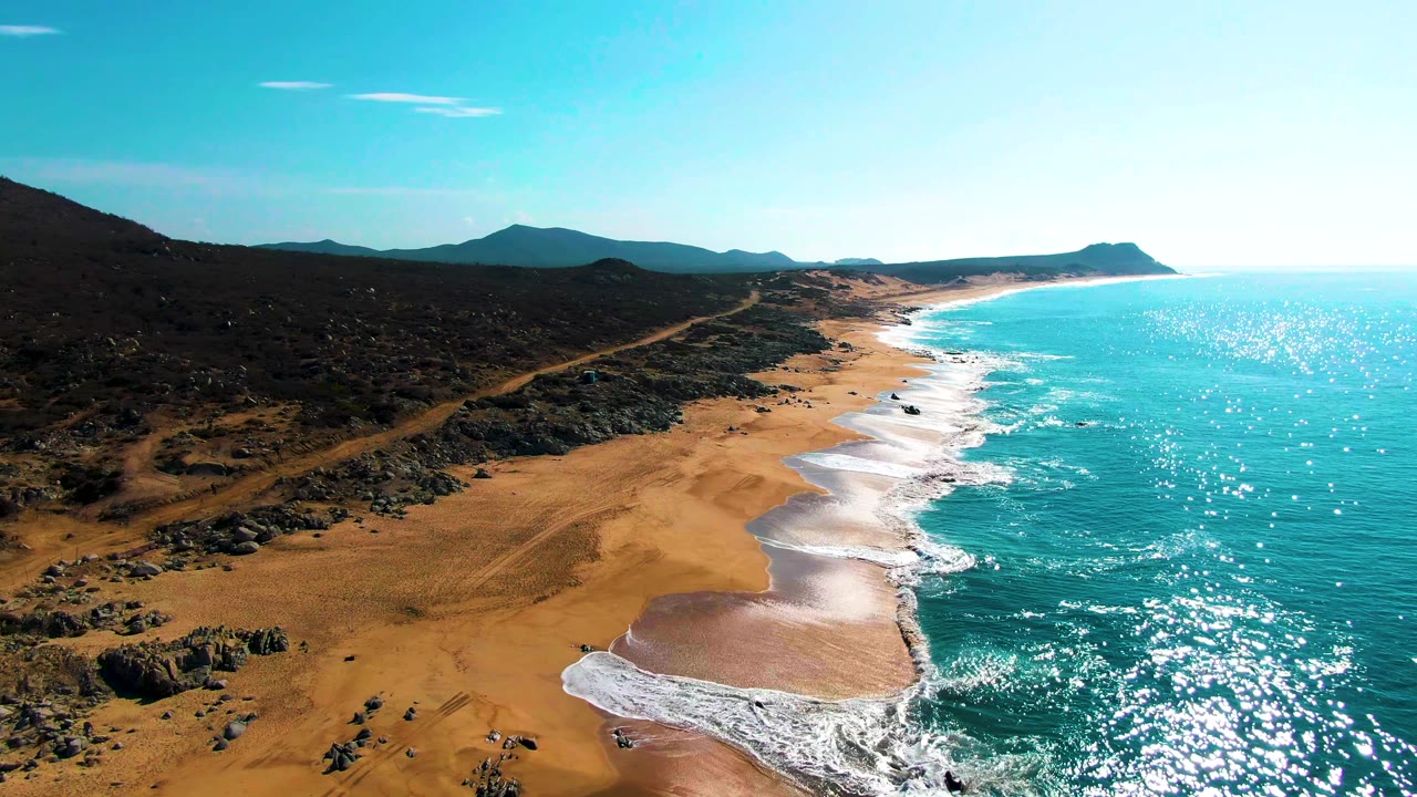 Bright sun reflections cover the turquoise ocean beside Buzzards Beach, with pale surf edging the sand, dark inland rocks and scrub, low hills in the distance, and no visible people.