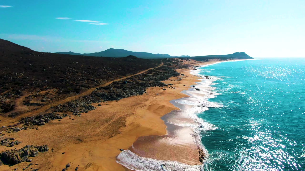 Aerial coastline with foamy waves, wide sand, rocky scrubland, and a dirt road inland.
