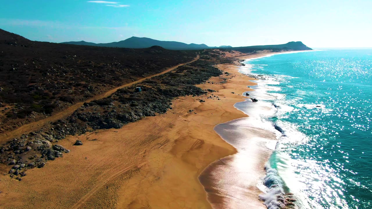 Aerial view of sandy beach, rocky coast, dirt road, turquoise ocean, and distant hills.