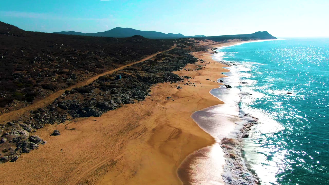 Drone view along Buzzards Beach with the road receding through dark rocky terrain, waves curling onto the sand, and no visible people.