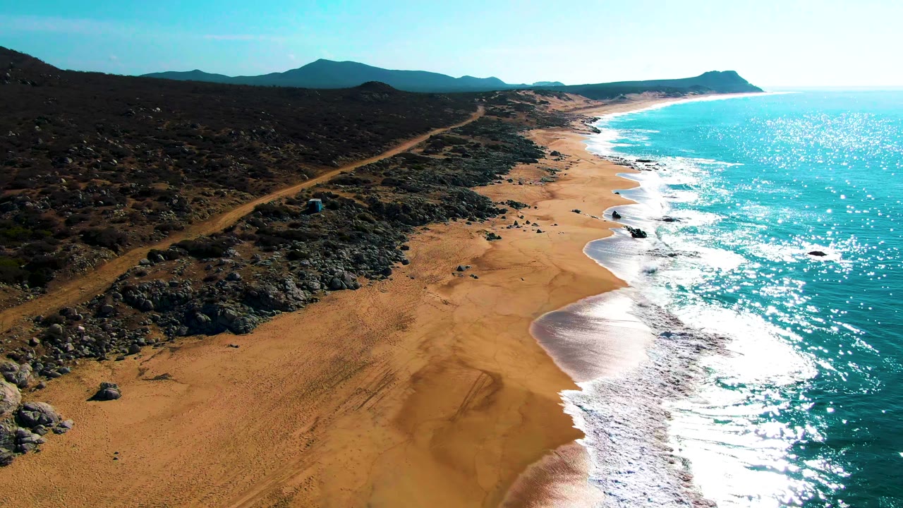 High aerial frame over Buzzards Beach showing stronger sun sparkle on the turquoise water, a curving wet shoreline, rocky inland ground, and no visible people.