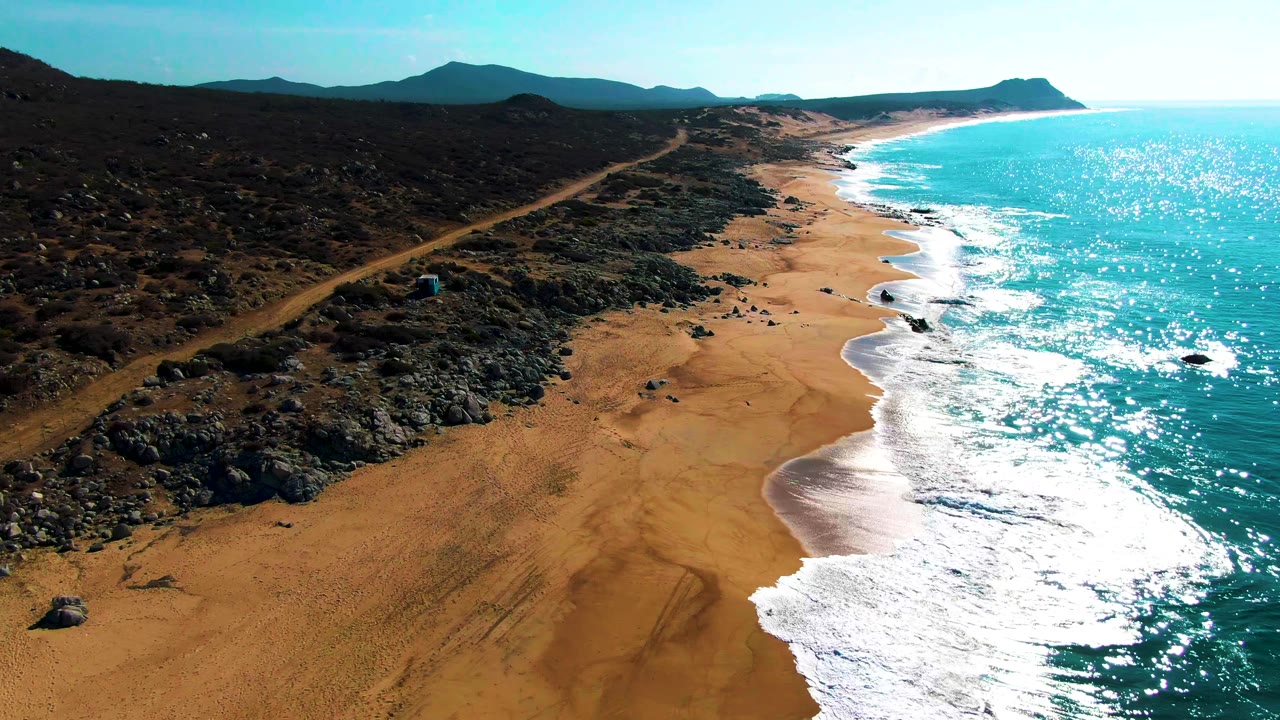 Later aerial frame at Buzzards Beach with the dirt road and rocky scrub more centered, the sandy beach widening below, and no visible people.