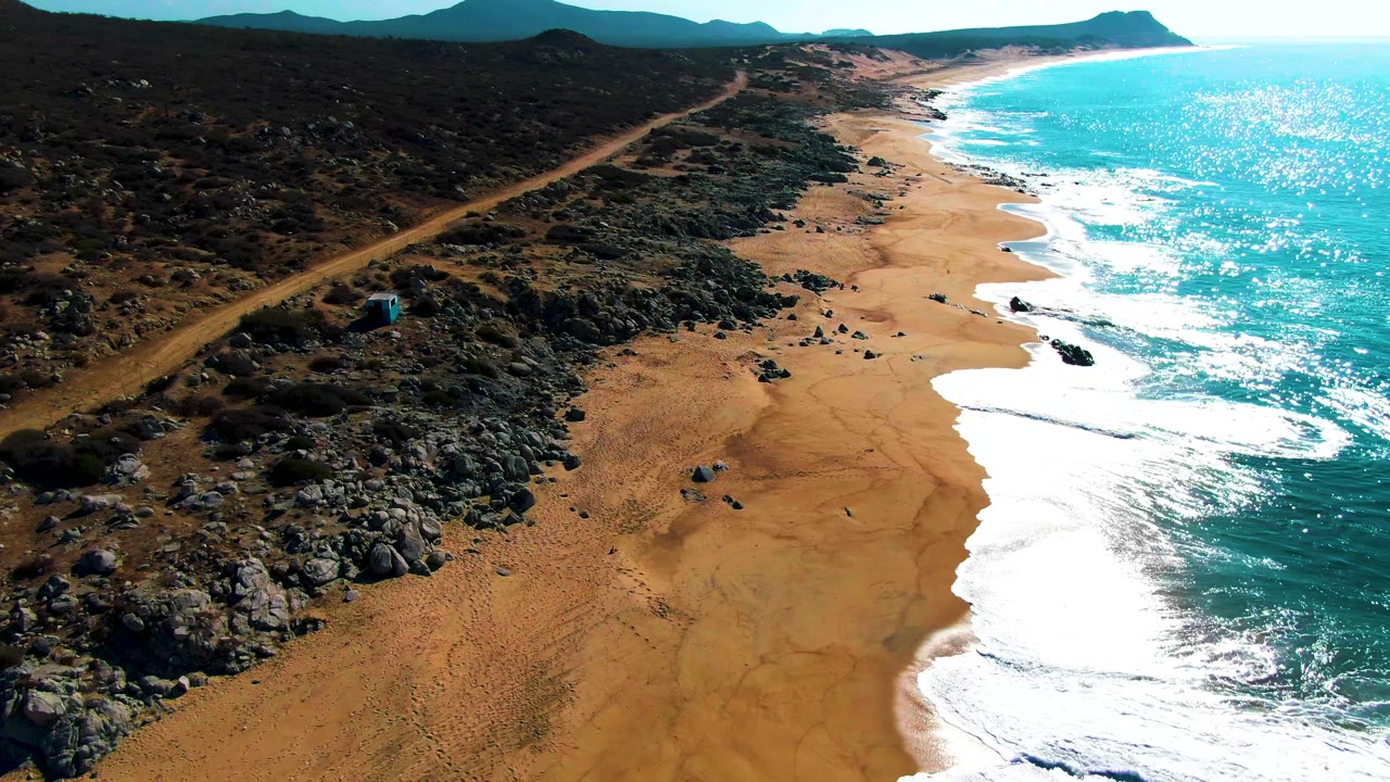 Aerial view of sandy beach, rocky desert, dirt road, turquoise ocean, and distant headland.