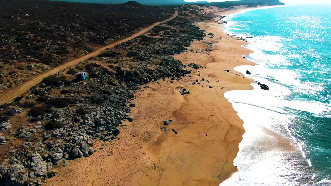 Aerial frame over Punta Buzzards showing the dirt road and small blue structure more clearly beside rocky desert terrain, with sandy beach and white surf curving along the turquoise ocean and no visible people.