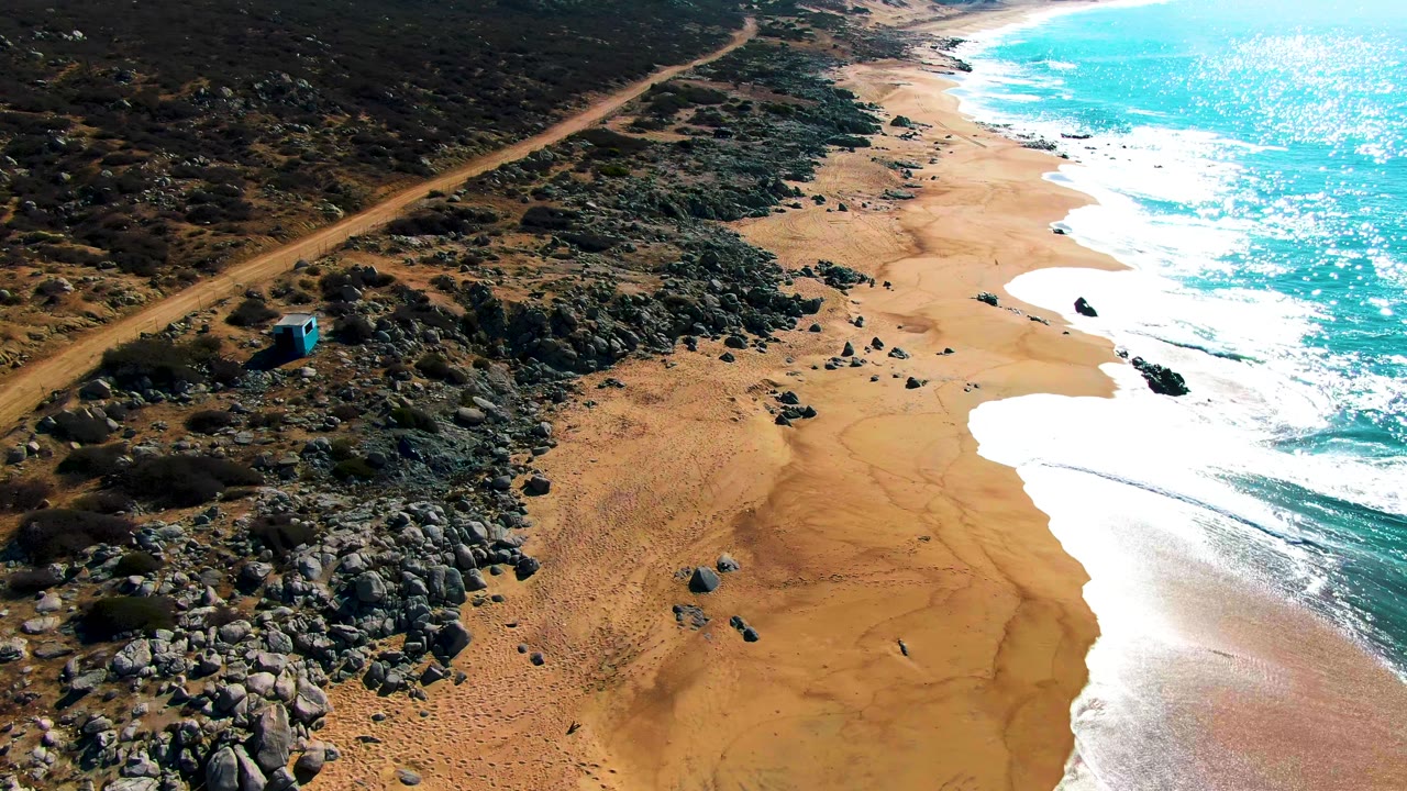 Closer aerial view at Punta Buzzards with the blue structure near the rocky scrub on the left, tan sand and scattered boulders below, white foam on the right edge, and no visible people.