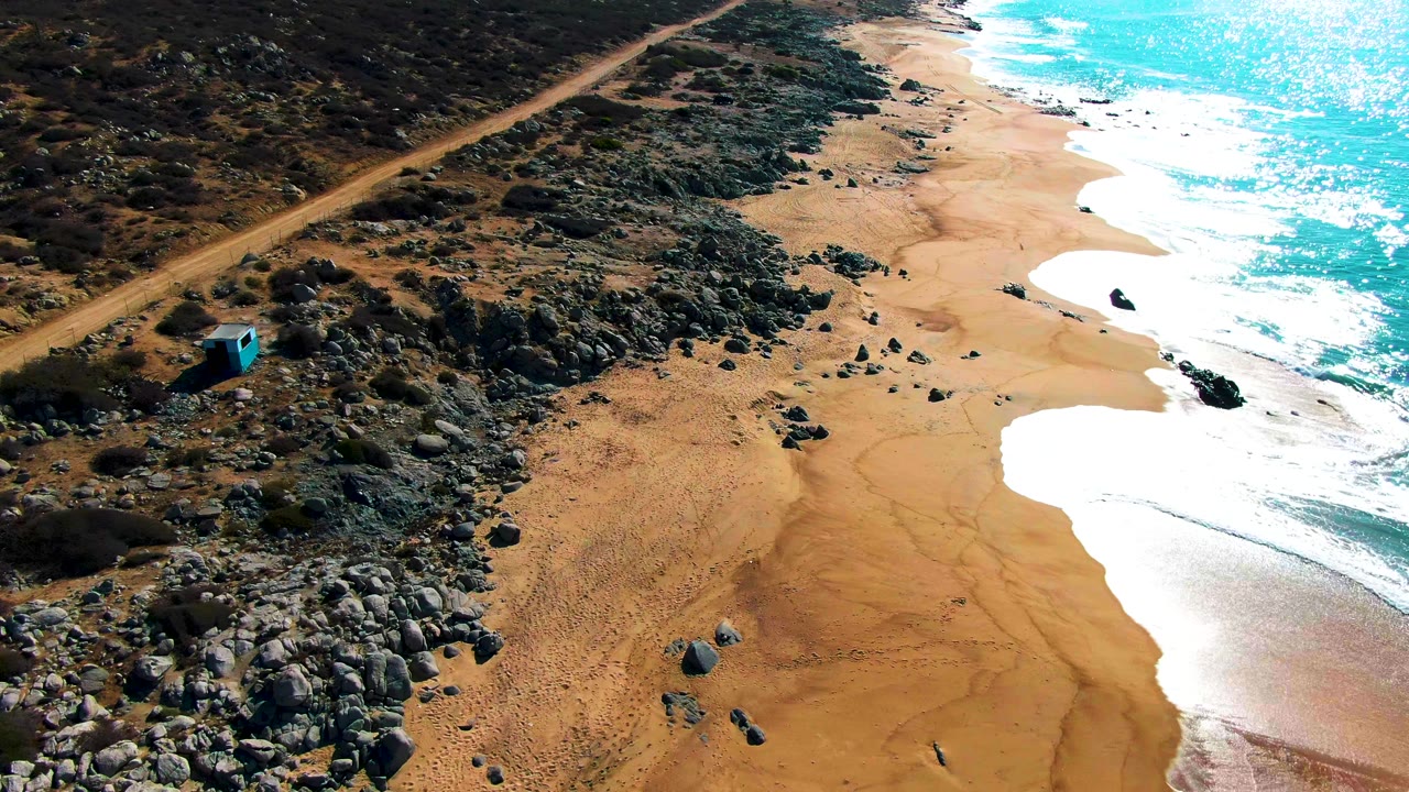 Aerial frame at Punta Buzzards with the small blue structure low on the left, dirt road crossing the upper left, rocky scrub and boulders through the center, surf washing the sandy shore, and no visible people.