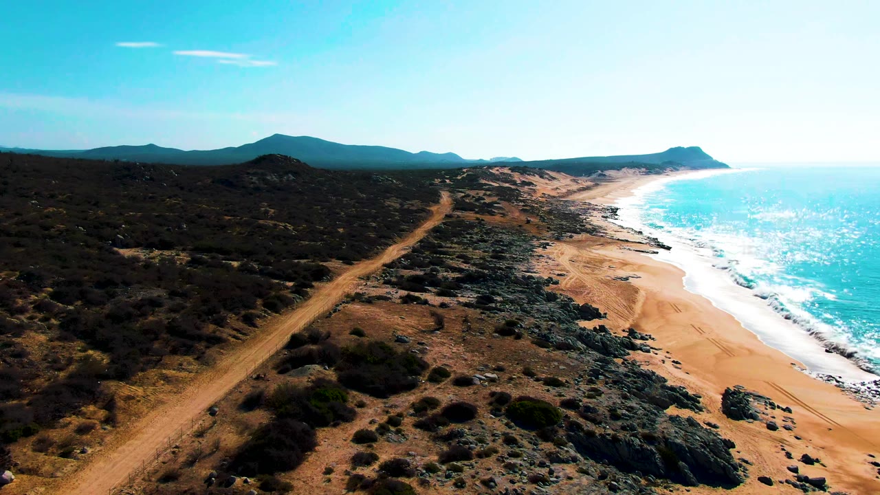 Aerial frame over Punta Buzzards, Baja California Sur, with a dirt road on the left, rocky scrub in the center, blue ocean on the right, distant hills, and no visible people.