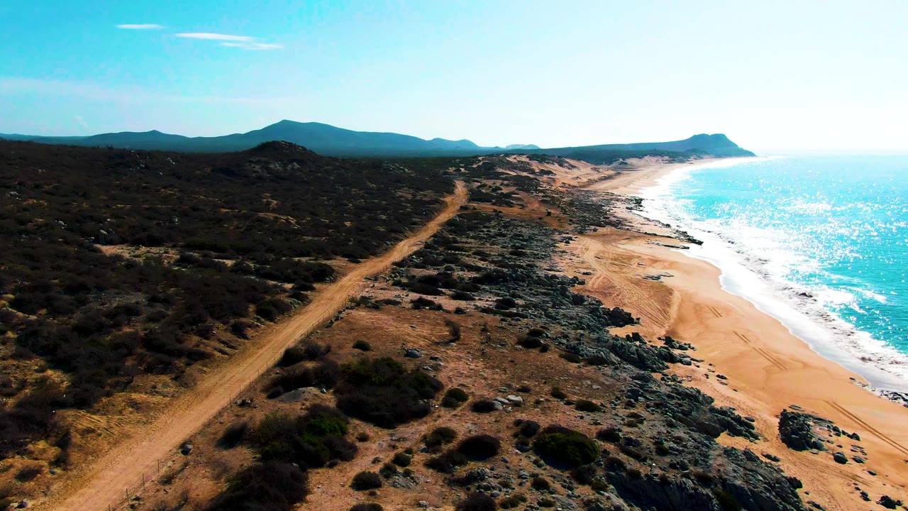 Aerial frame over Punta Buzzards showing the tan road closer in the foreground, dark coastal scrub, a narrow sandy beach, bright ocean water, distant mountains, and no visible people.